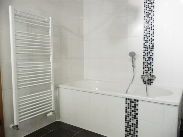 Weiße Badewanne mit schwarz-weißen Mosaikstreifen und schmalem Handtuchheizkörper an heller Fliesenwand