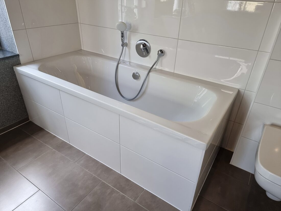 Weiße Badewanne mit Handbrause und beigen Fliesen, daneben WC auf grauem Boden
