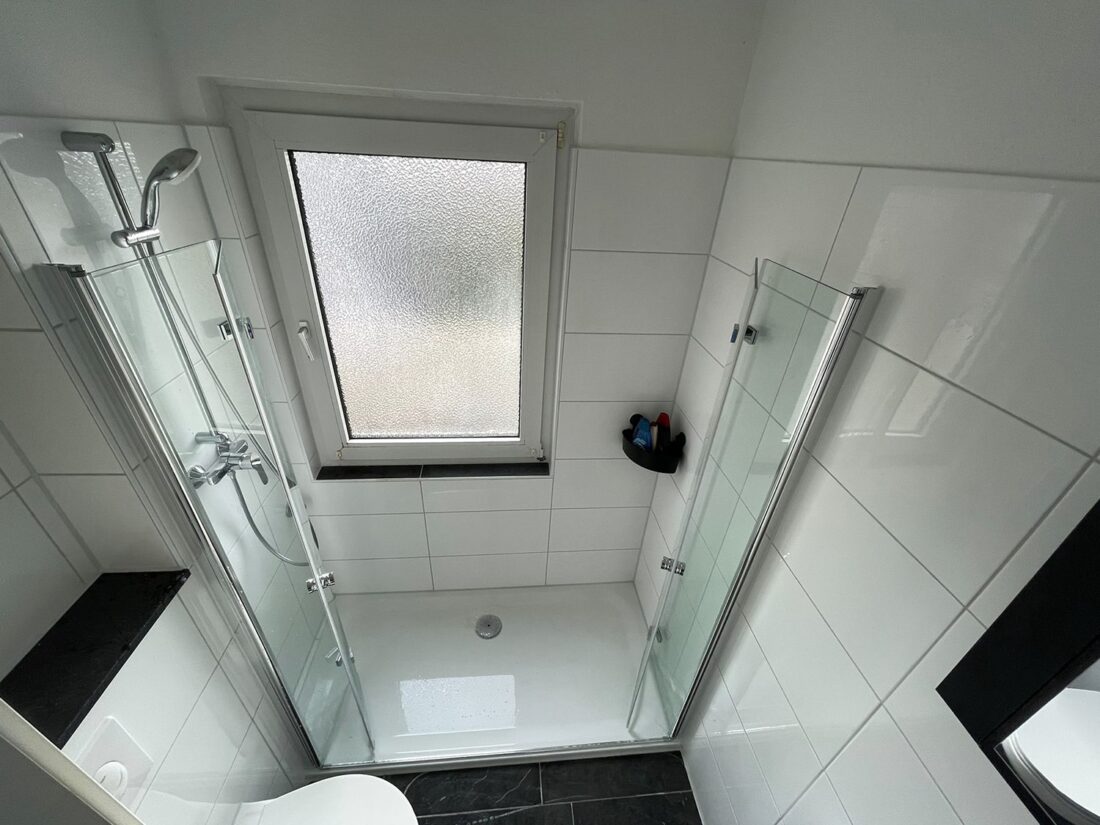 Bodengleiche Dusche mit Glasfaltwand und Fenster, weiße Fliesen, schwarzer Steinboden – Badsanierung in Essen