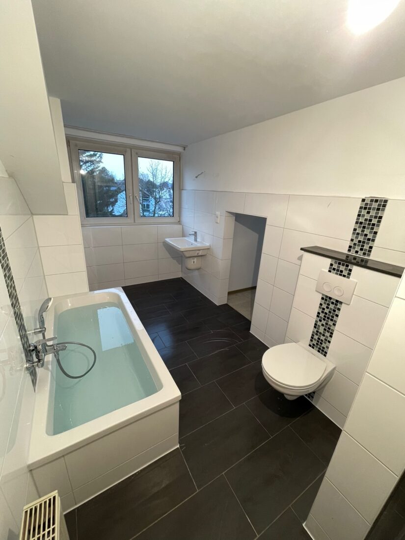 Frisch saniertes Bad mit Badewanne, Wand-WC und Fenster, dunkler Bodenfliese und Mosaikakzent